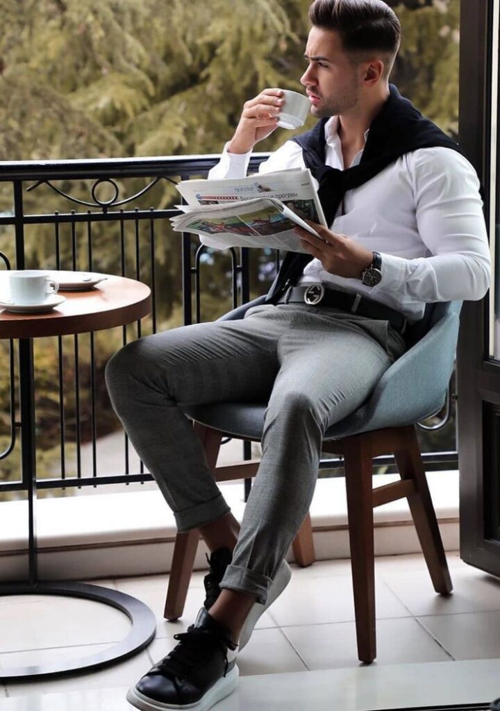 A man in a white shirt and gray pants sits on a balcony chair, reading a newspaper and sipping coffee, with a scenic view of trees in the background.