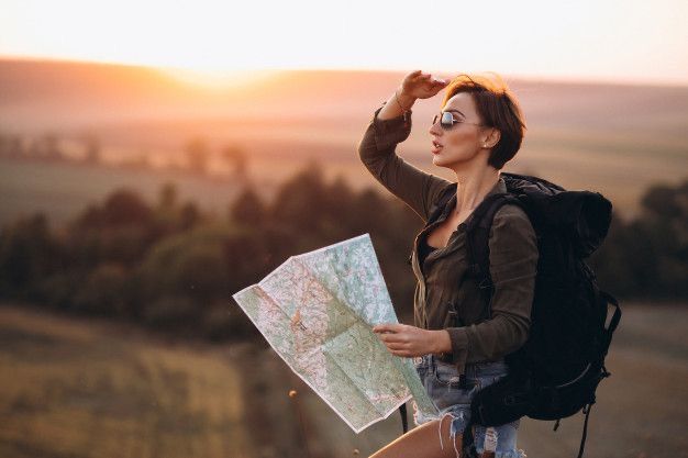 A female traveler with a backpack holding a map and looking towards the horizon during sunset in a scenic outdoor location.